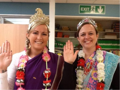 Two women wearing traditional clothing and floral garlands, smiling and raising their hands.